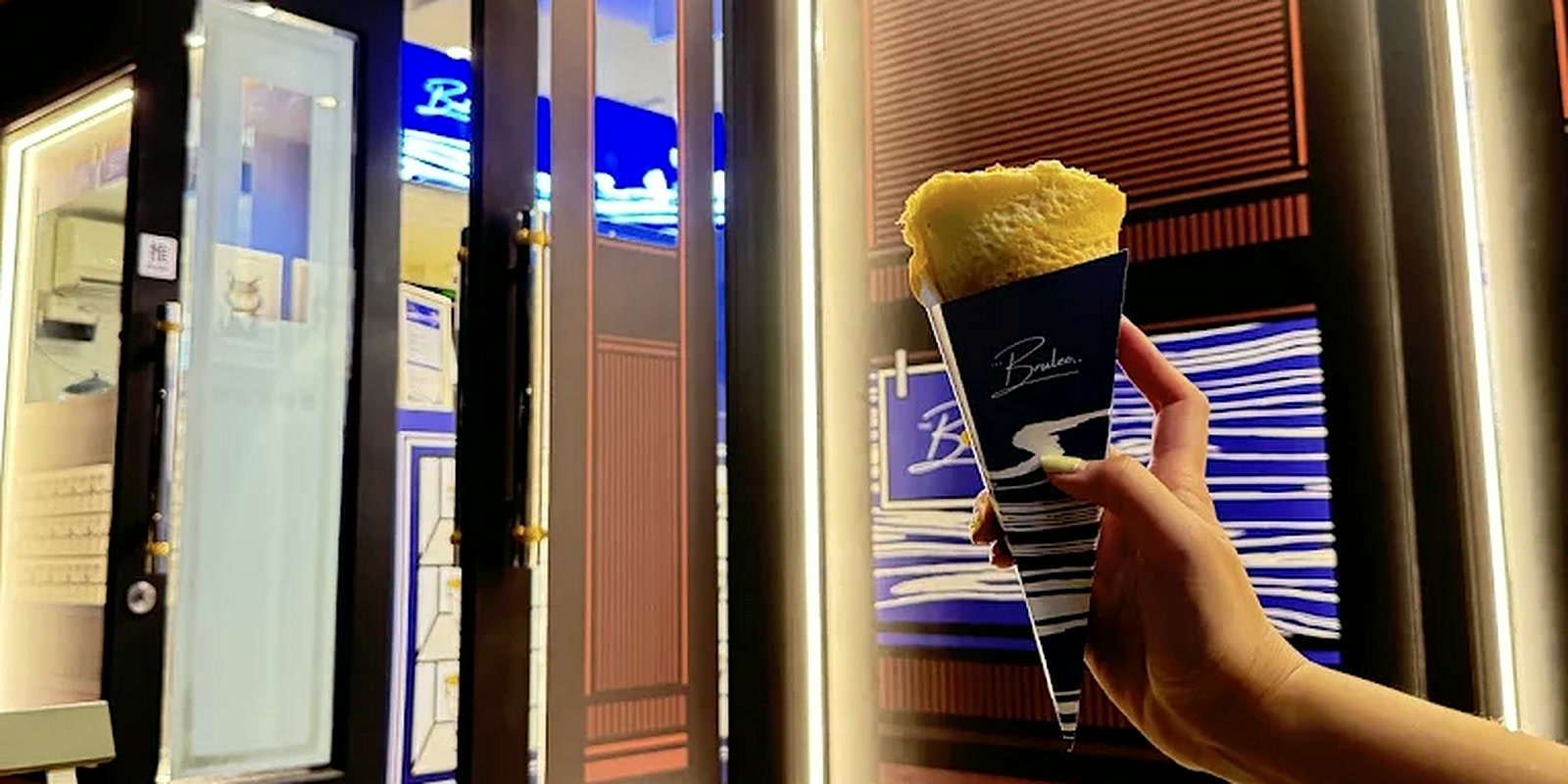 A Brulee crepe brulee in its branded cone, held up outside the lit shopfront