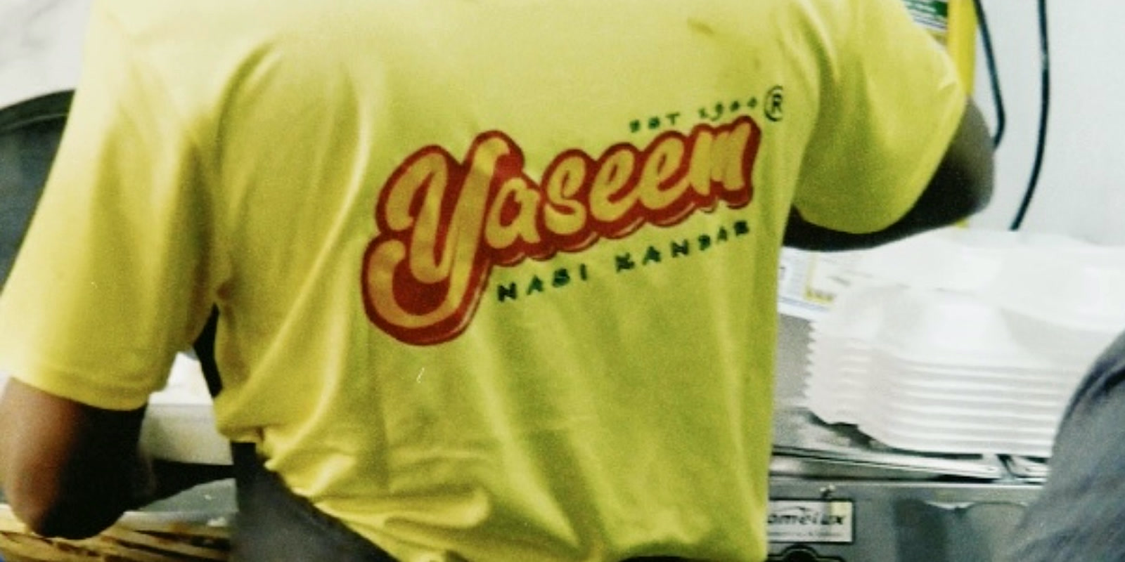 A Yaseem Nasi Kandar staff member working the counter, the brand logo on the back of the uniform